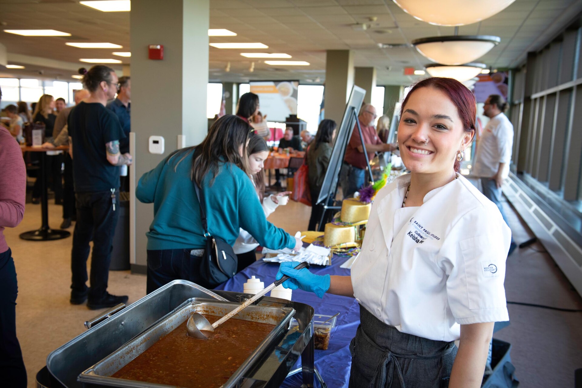 A student in a white chef coat and gray apron stands next to a serving station with a large pan of chili and a ladle. Behind the station, attendees are interacting with other food booths in a bright room with large windows.