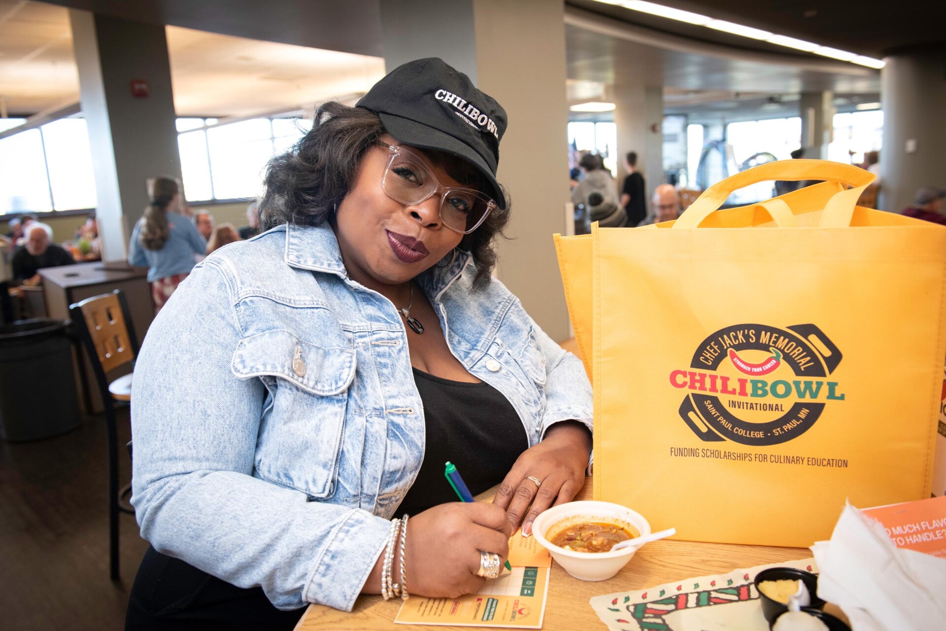 Person wearing a denim jacket and a black cap labeled “CHILBOWL” writes on a scorecard at a table. A bright yellow tote bag with the “Chef Jack’s Memorial Chili Bowl Invitational” logo is placed next to a bowl of chili.