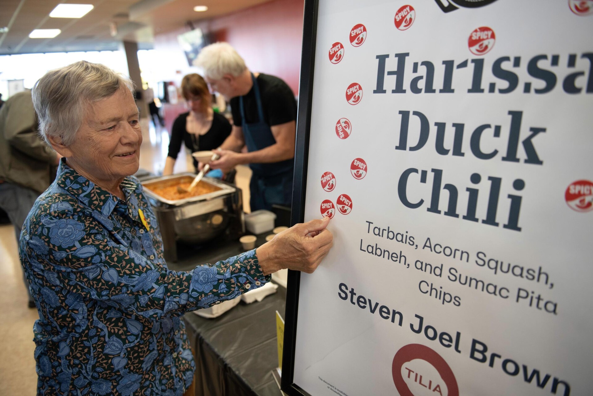 Person in a patterned blue shirt applies a sticker to a sign that reads “Harissa Duck Chili” with listed ingredients including tarbais, acorn squash, labneh, and sumac pita chips. Behind the sign, two people serve chili from a metal pan.