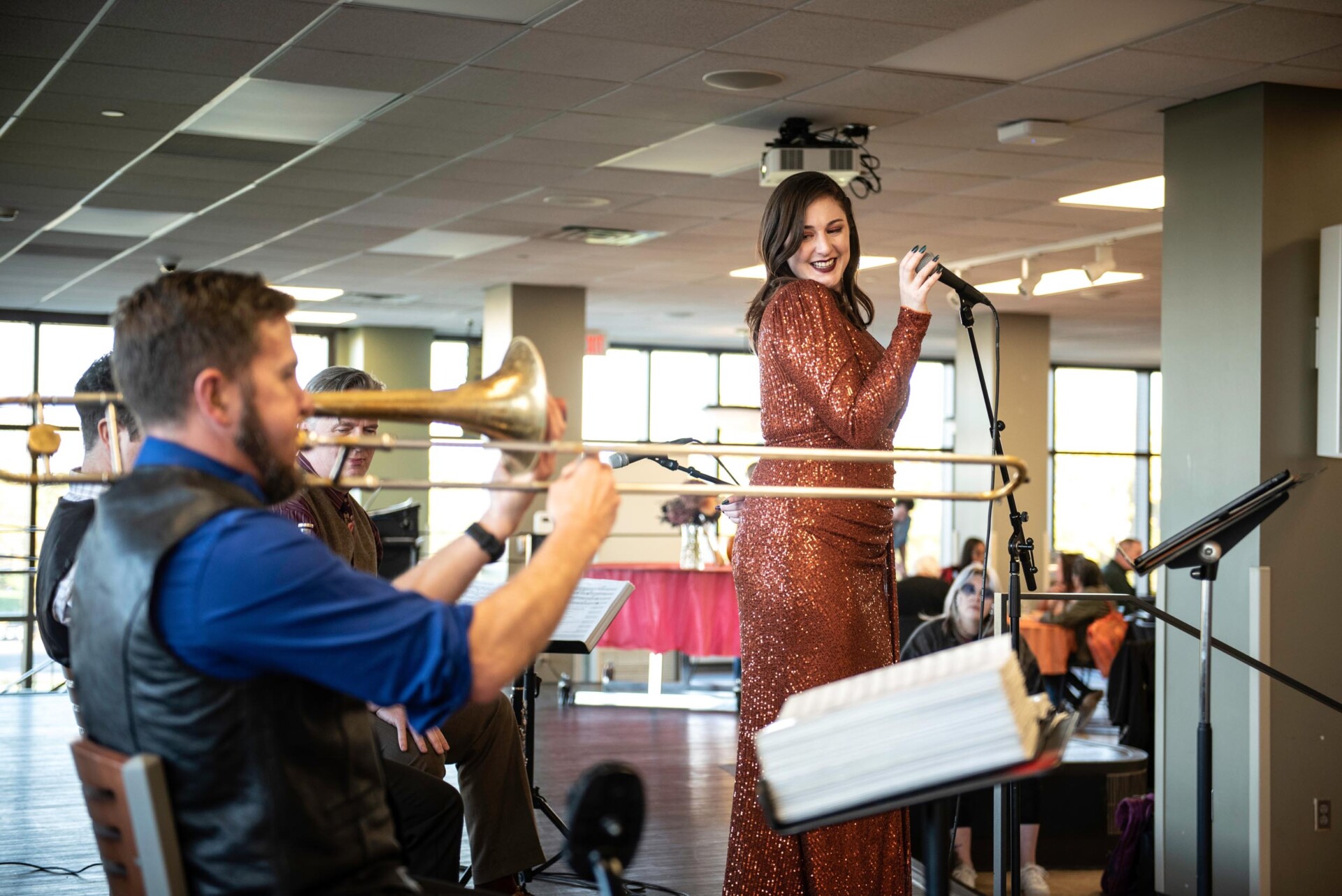 A musician in a blue shirt plays a trombone while another performer in a sparkling copper-colored gown sings into a microphone. Music stands and microphones are visible in the foreground, with large windows in the background.
