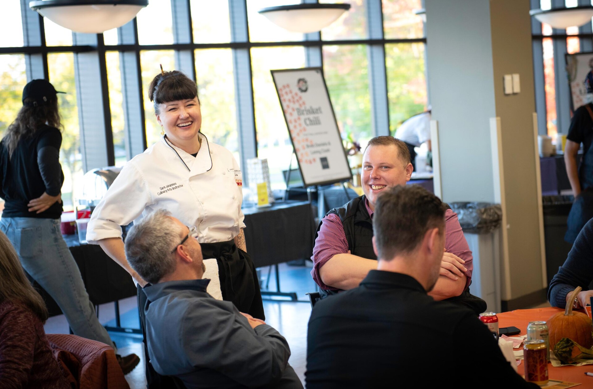 Sara Johnannes in a white chef coat stands near a table where three seated individuals are engaged in conversation