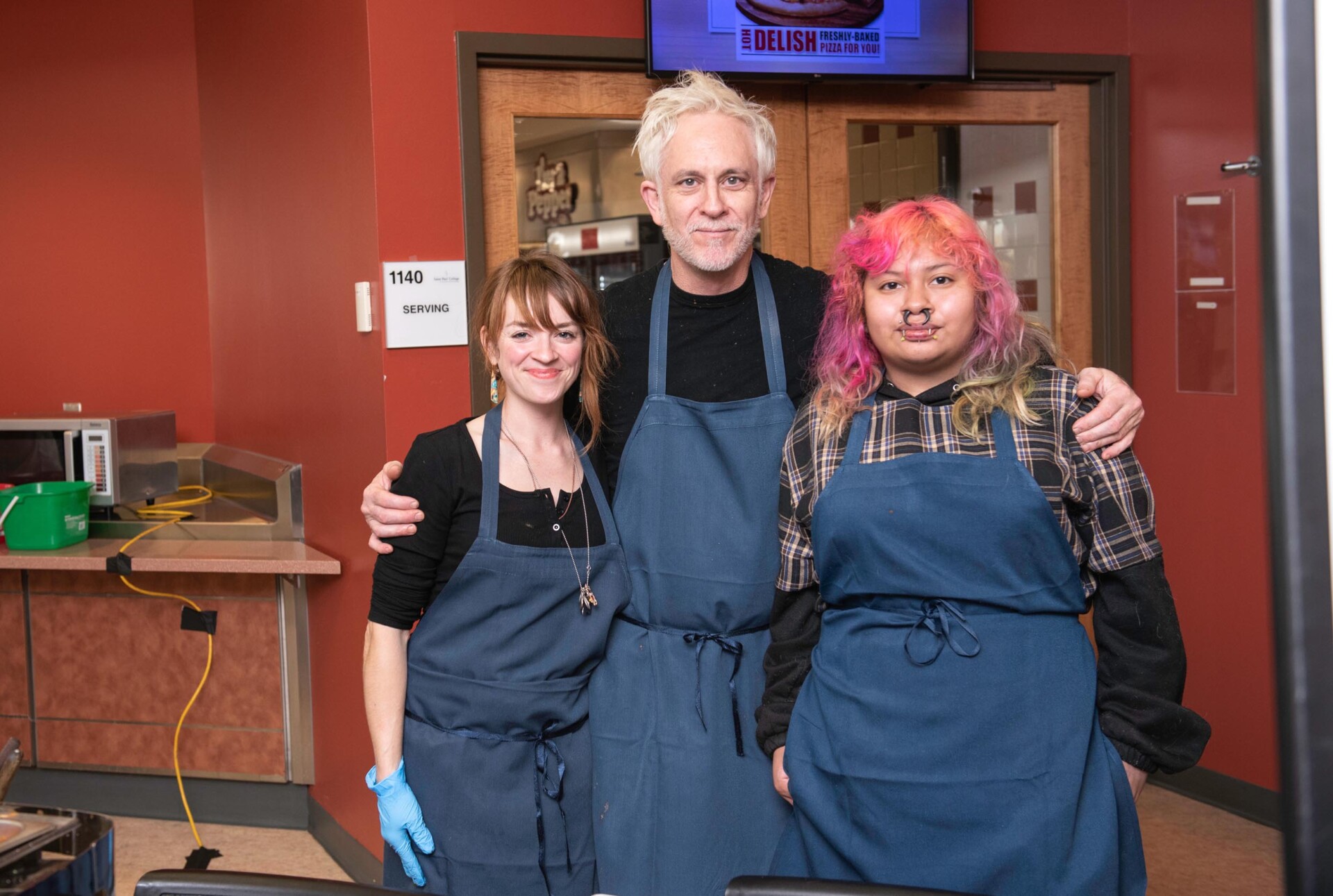 Three people wearing dark aprons stand together in a kitchen area with red walls. One person has an arm around the others, and a microwave and utensils are visible in the background.