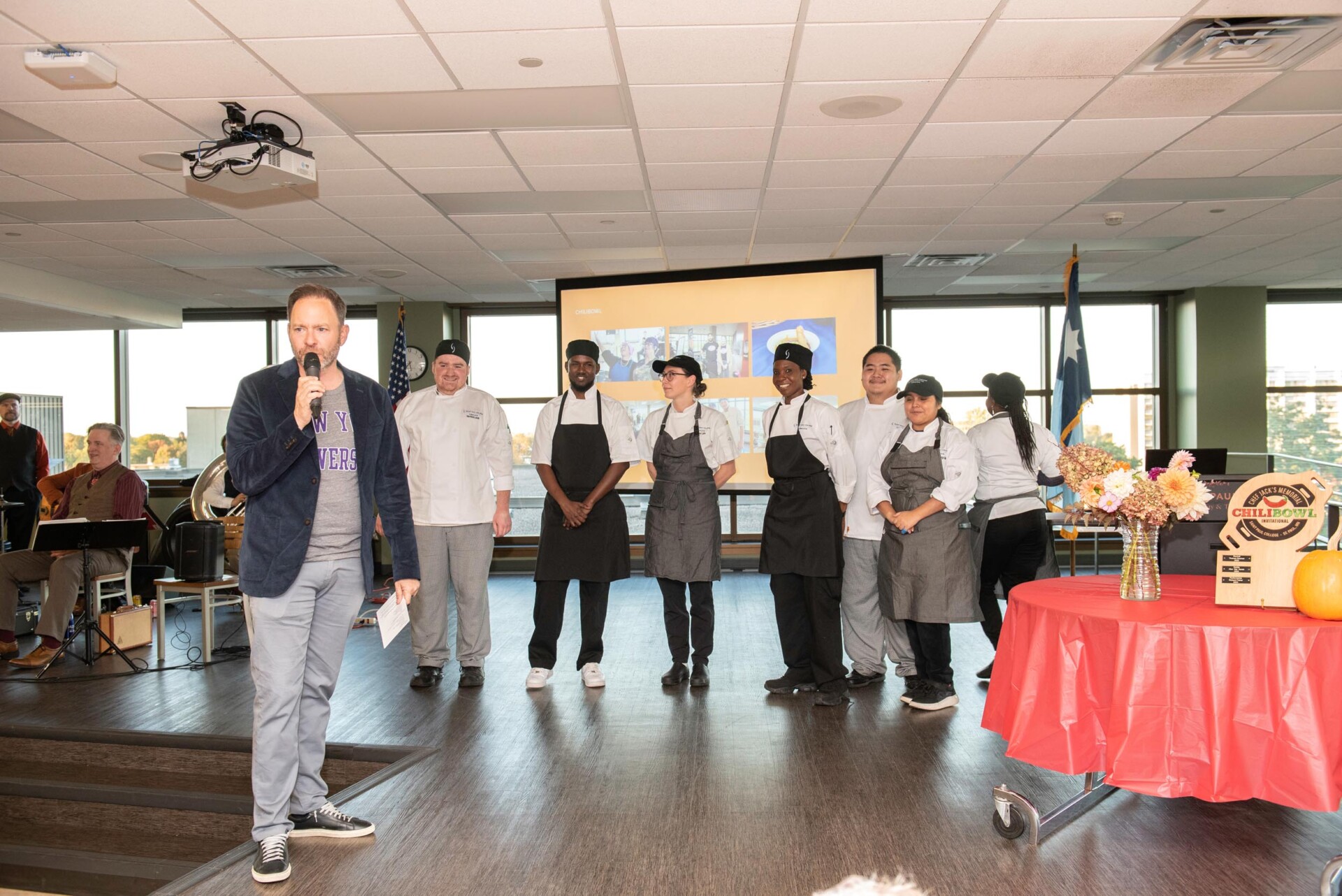 A group of culinary students and staff stand on a stage wearing white chef coats and gray aprons. Jason DeRusha with a microphone stands to the left, and a table with flowers and a Chili Bowl award plaque is on the right. A presentation screen and American flag are visible in the background.