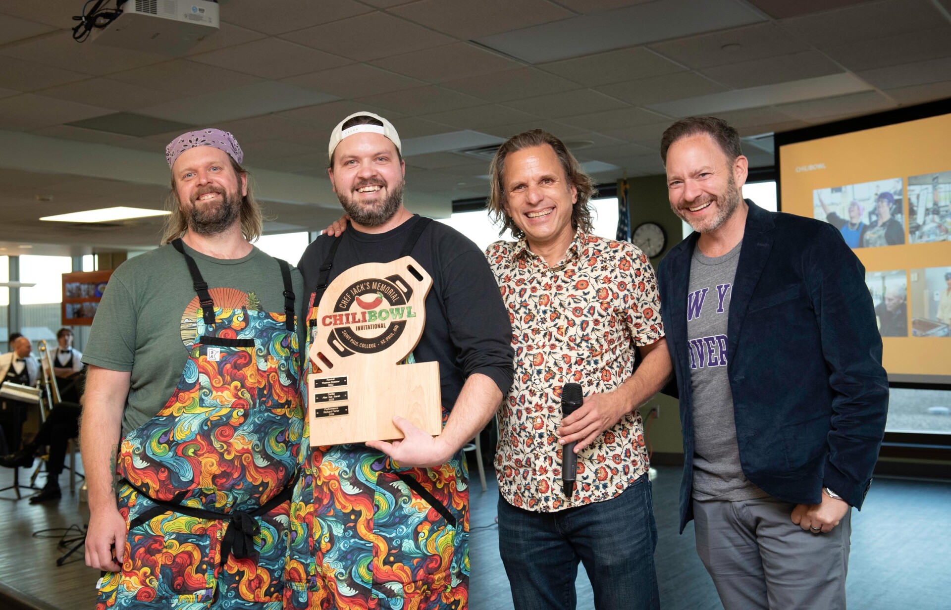 Four people stand together indoors, with one person holding a wooden award plaque that reads “Chili Bowl Invitational.” Two individuals wear colorful aprons with swirling rainbow patterns, and a presentation screen is visible in the background.