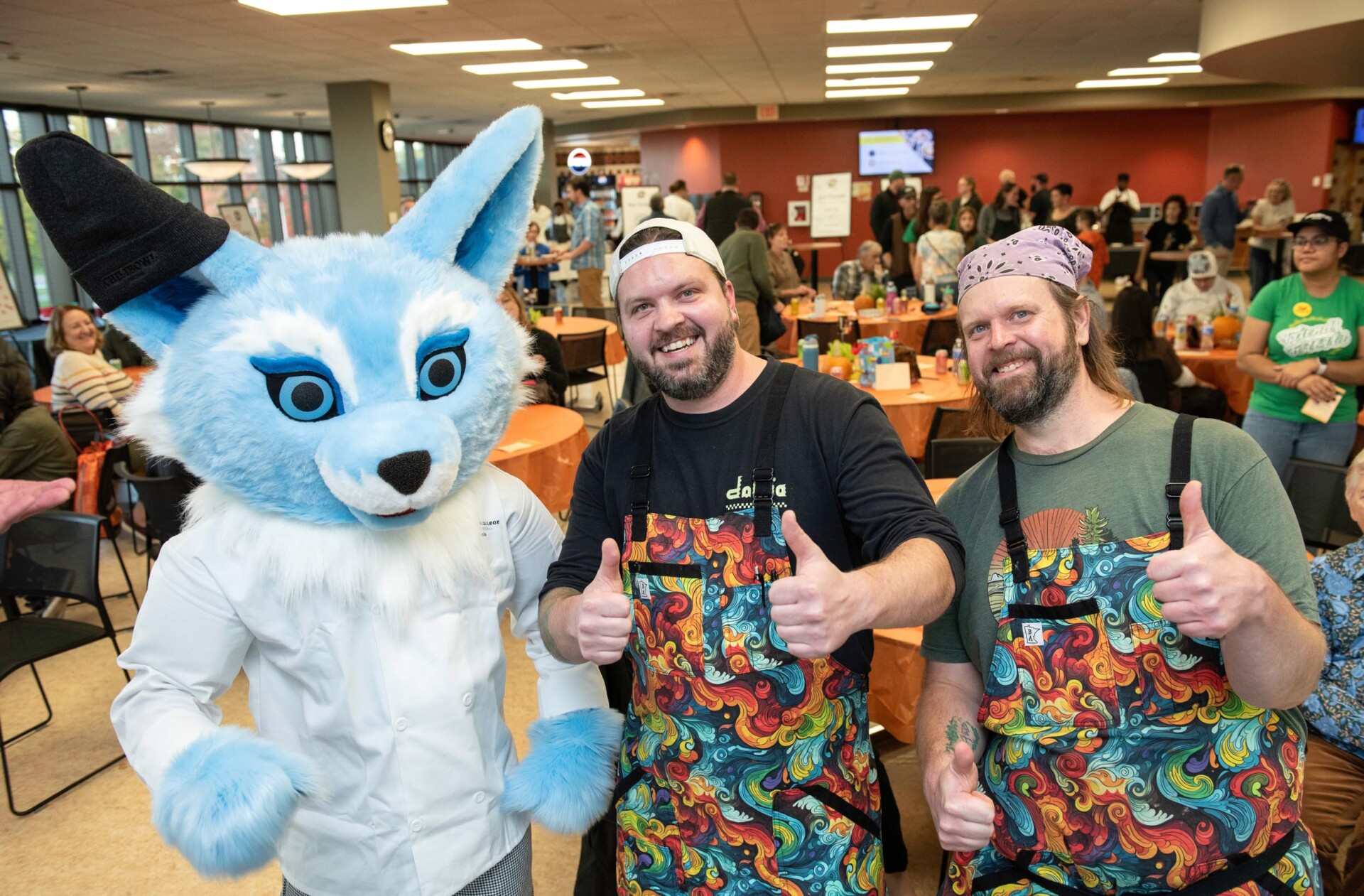 Sky the fox mascot wearing a white chef coat poses with two individuals giving thumbs up. Both people wear matching aprons with colorful swirling designs. The background shows a crowded event space with orange tablecloths and attendees.