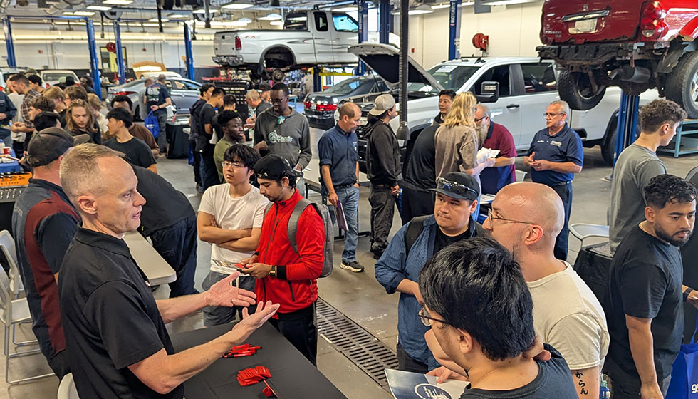 A bustling automotive workshop during a career fair or open house event. A person in a black shirt speaks to a group of attendees holding pamphlets, while others work on cars elevated on hydraulic lifts in the background.