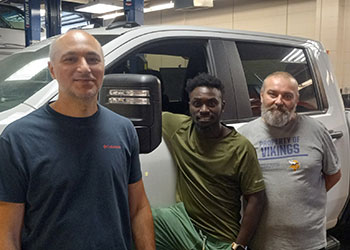 Three individuals standing in front of a white truck inside a garage. They are dressed in casual t-shirts—dark blue, olive green, and light grey with 'Property of Vikings' text and a yellow bird graphic. The truck's side mirror and window are visible, with garage equipment in the background.