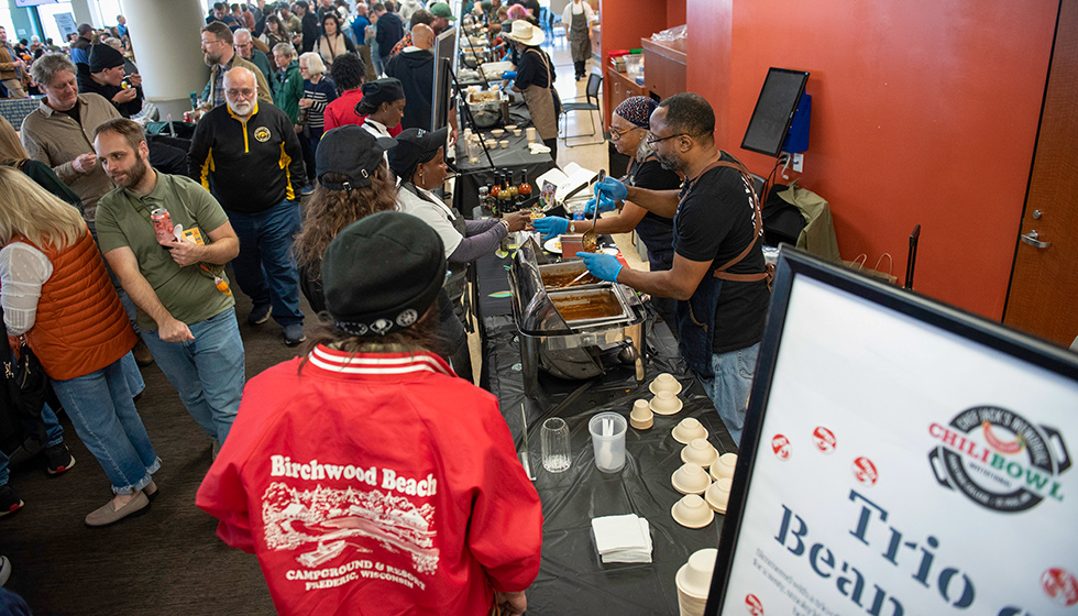 A busy chili-tasting event with a long serving table covered in black cloth. Two people wearing aprons and gloves serve chili from large pans while attendees wait in line. The background shows a crowded room with multiple food stations.