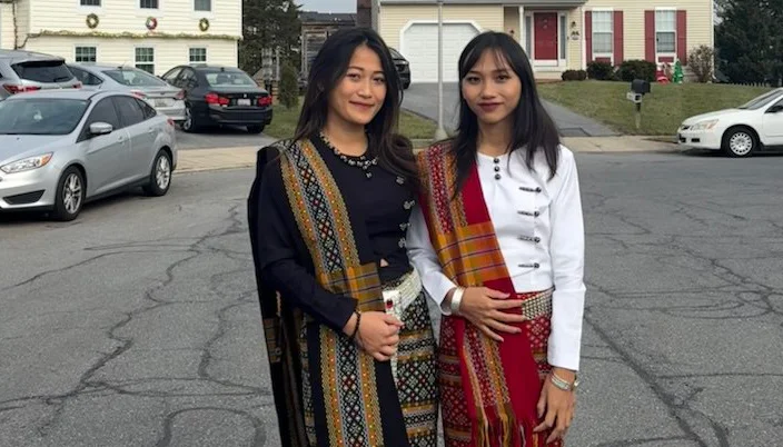 Two individuals standing on a residential street wearing traditional woven garments with intricate patterns and colorful sashes draped over their shoulders. One person is dressed in a black outfit with a multicolored patterned sash, while the other wears a white top and red skirt with a similar patterned sash. Behind them are parked cars and houses with light-colored siding and red shutters.