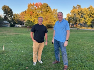 Tom Leiskau and Tom Hillstead standing on a grassy field with autumn-colored trees and houses in the background. Both are smiling and facing the camera.
