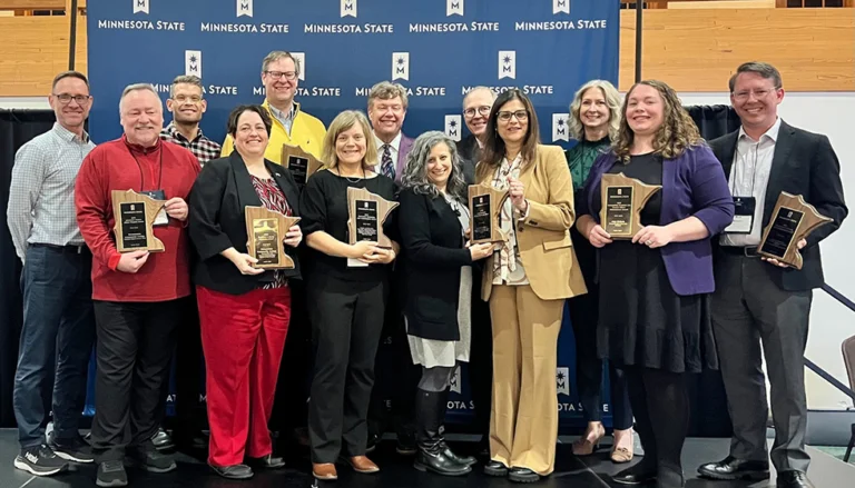 A group of people standing in front of a blue backdrop with the Minnesota State logo repeated across it. Each person is holding a wooden plaque shaped like the state of Minnesota, indicating an award ceremony setting.
