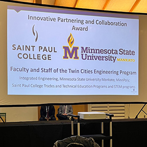 A large projection screen displays the “Innovative Partnering and Collaboration Award” title. Below it are the logos for Saint Paul College and Minnesota State University Mankato. The text recognizes the faculty and staff of the Twin Cities Engineering Program, mentioning Integrated Engineering, Minnesota State University Mankato, MinnePoly, Saint Paul College Trades and Technical Education Programs, and STEM programs. A table and chairs are visible in front of the screen.