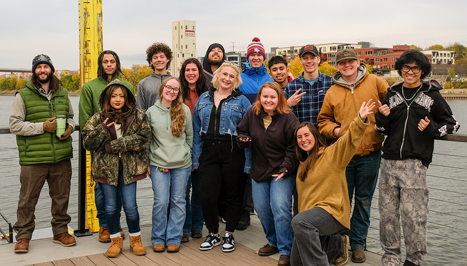 A group of students standing on a barge in the Mississippi River, posing for a photo. A tall yellow river gauge is visible on the left, and buildings with autumn-colored trees are in the background.