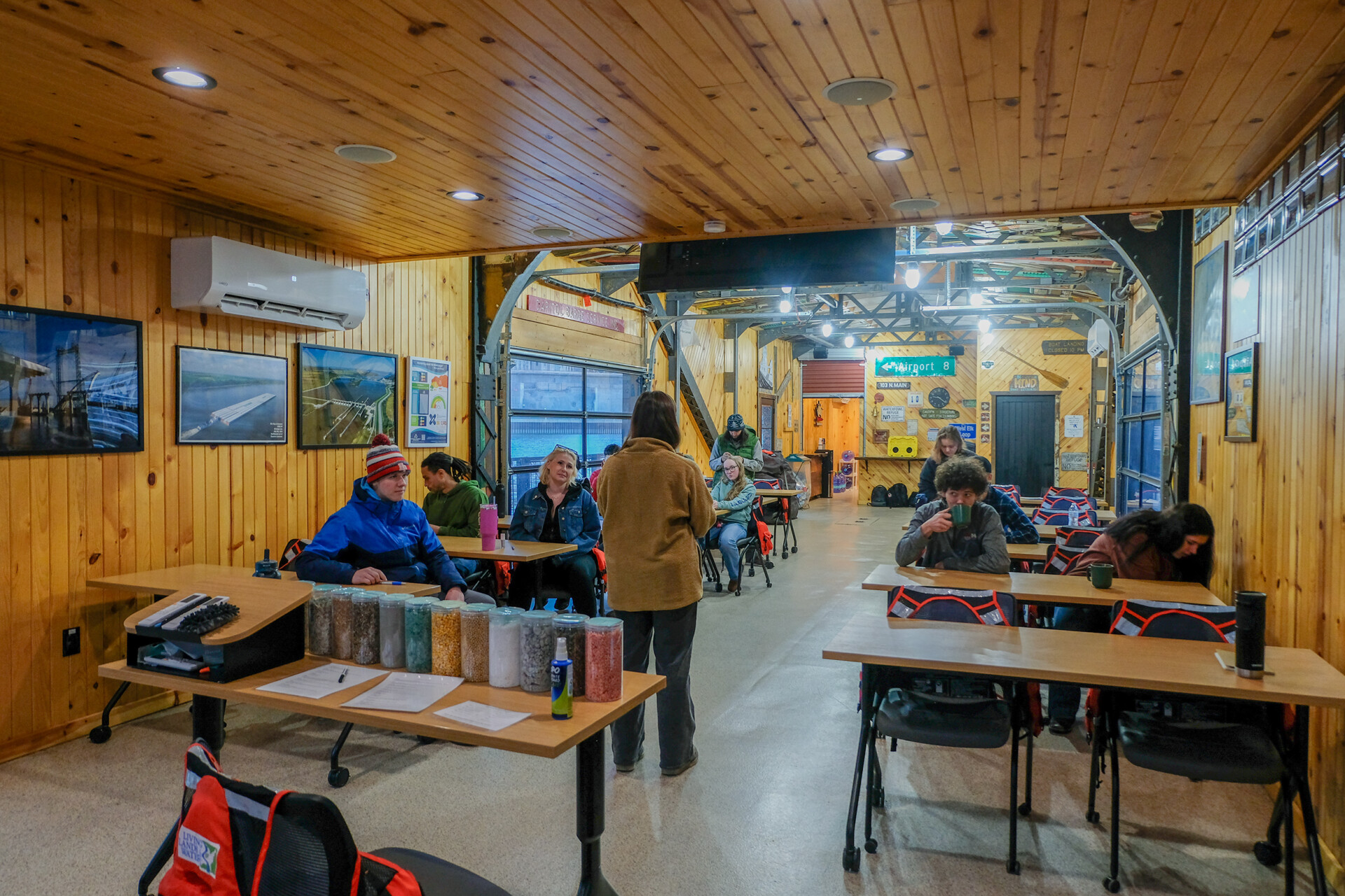 An indoor classroom or wooden walls and ceiling. Several people are seated at tables while someone stands at the front speaking. The room has framed photos on the walls and supplies on the tables.