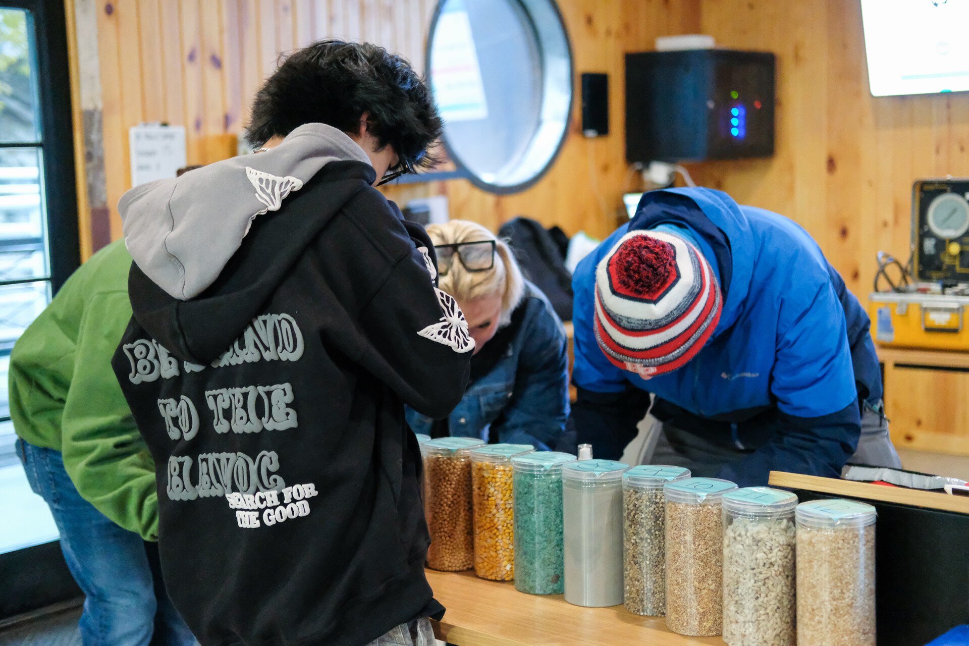 Several students gathered around a table inside a wooden-paneled room, examining clear containers filled with various grains and seeds arranged in a row. A round window and electronic equipment are visible in the background.