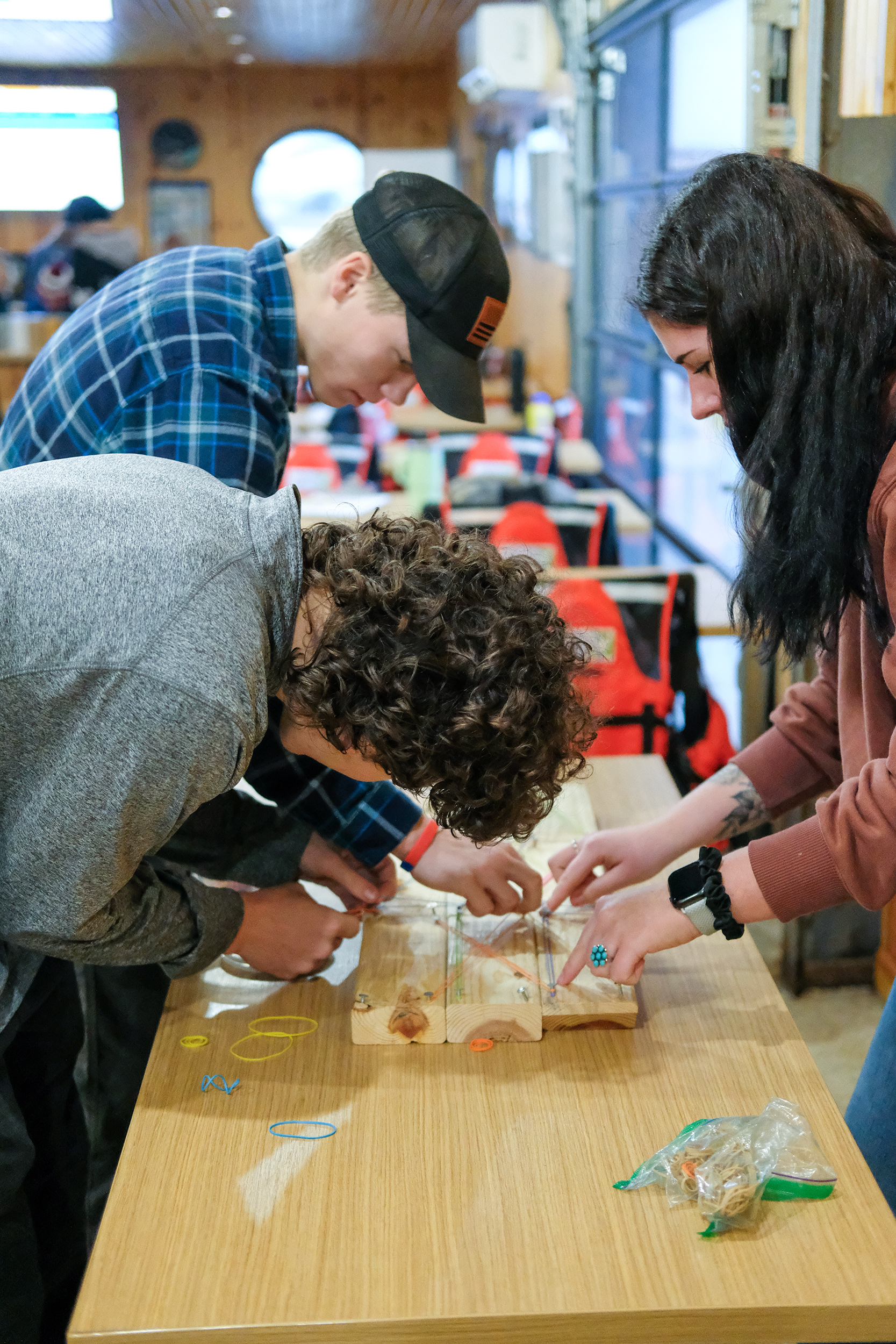 Three students leaning over a wooden table, working on a hands-on activity involving small objects, rubber bands, and wooden pieces. Red chairs and large windows are visible in the background.