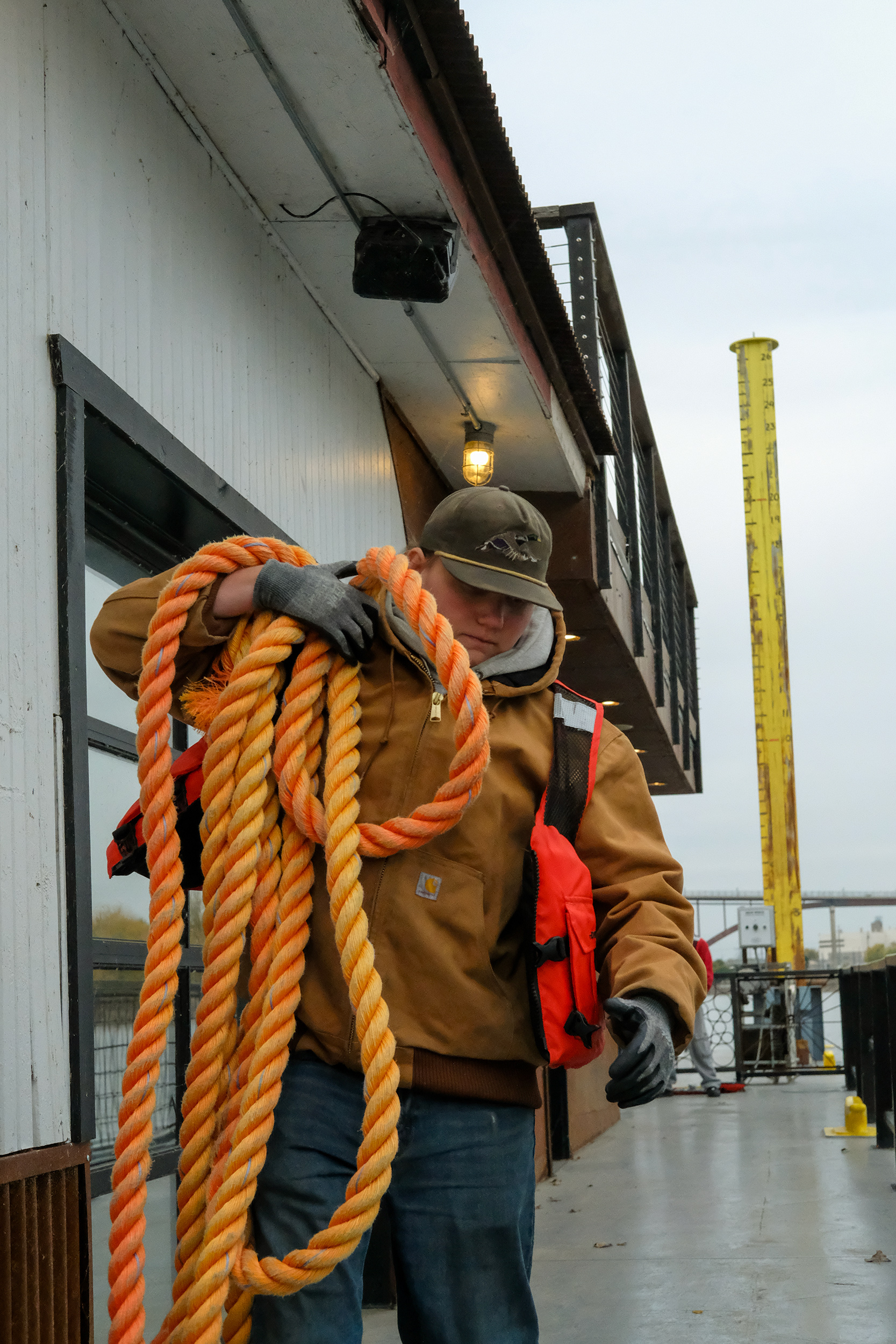A student wearing a brown jacket and gloves carrying a large coil of thick orange rope on an outdoor deck. The deck has metal railings and a building structure with an upper balcony.