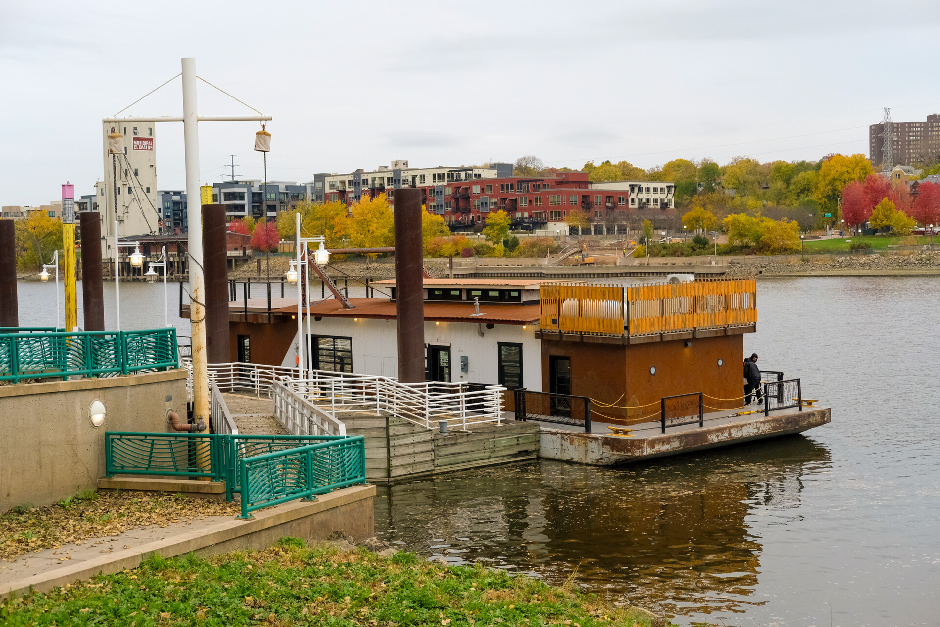A floating barge with a wooden deck and railings moored along a riverbank. The background shows autumn trees, modern buildings, and a cloudy sky.