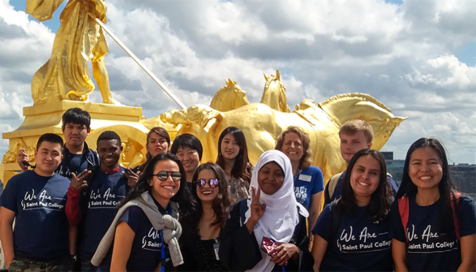 Group of students wearing matching navy blue T-shirts with ‘We Are Saint Paul College’ printed on them, standing in front of a large golden statue featuring a figure holding a staff and a horse at the Minnesota State Capitol, under a partly cloudy sky