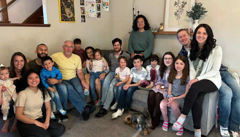 A group of adults and children sit and stand together in a living room, smiling at the camera; a small dog is on the floor in front of them.