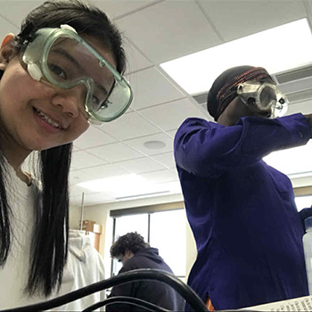 Two students in protective goggles work in a science lab; one is facing the camera and smiling, the other is focused on an experiment.