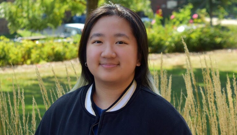 Cabee Thao wearing a dark jacket with a white-trimmed collar standing outdoors in a landscaped area with tall ornamental grasses and green foliage in the background.