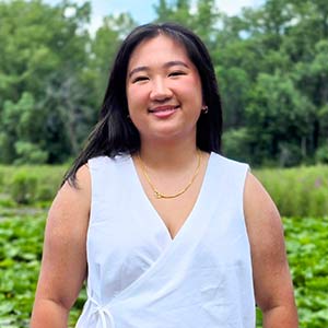 Cabee Thao wearing a sleeveless white wrap-style top with a gold necklace, standing outdoors in front of a lush green background with trees and a pond filled with lily pads.