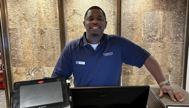Aaron Kirk standing behind a hotel front desk, wearing a navy Homewood Suites uniform shirt with a visible name badge. The desk has a monitor and a payment terminal, and the background features a textured wall with a warm, earthy tone.
