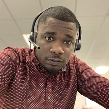 Aaron Kirk wearing a maroon shirt with a small white dotted pattern and over-ear headphones, seated indoors with a neutral office background featuring ceiling panels and lights.