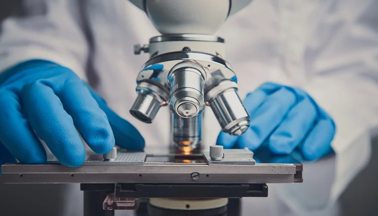 Close-up of a person wearing blue gloves adjusting a microscope slide under a silver compound microscope, with a white lab coat visible in the background, representing a scientific or laboratory setting.