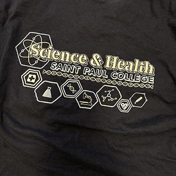 Black T-shirt featuring the words “Science & Health Saint Paul College” in bold lettering, surrounded by hexagonal icons representing science and healthcare fields, including a cross symbol, a laboratory flask, a DNA strand, molecular structures, and a heart with a pulse line.