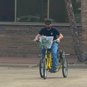 A person riding a custom-built yellow tricycle on a paved surface outdoors. The tricycle has a wide frame, two rear wheels, and a basket mounted on the front. A brick building and a tree trunk are visible in the background.