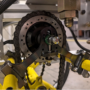 A close-up view of a mechanical assembly on a yellow frame, showing a chain drive connected to a sprocket and a metal bracket. The image highlights detailed components such as bolts, bearings, and part of a hydraulic or pneumatic fitting.