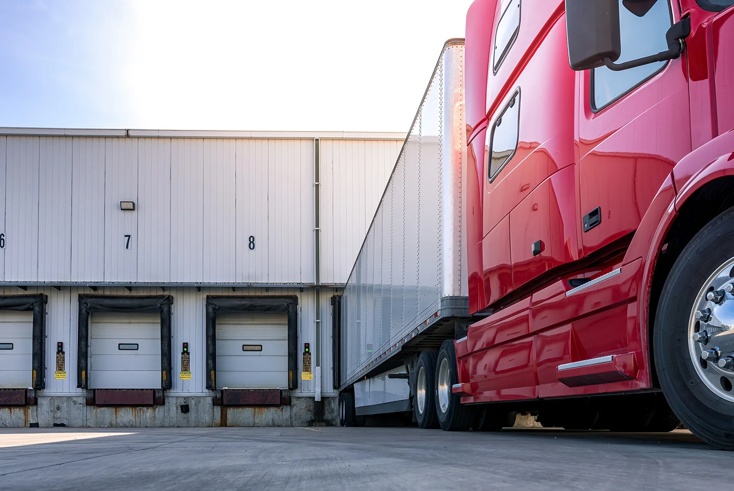 Red semi-truck parked at a loading dock with numbered bay doors on a warehouse building under clear daylight.