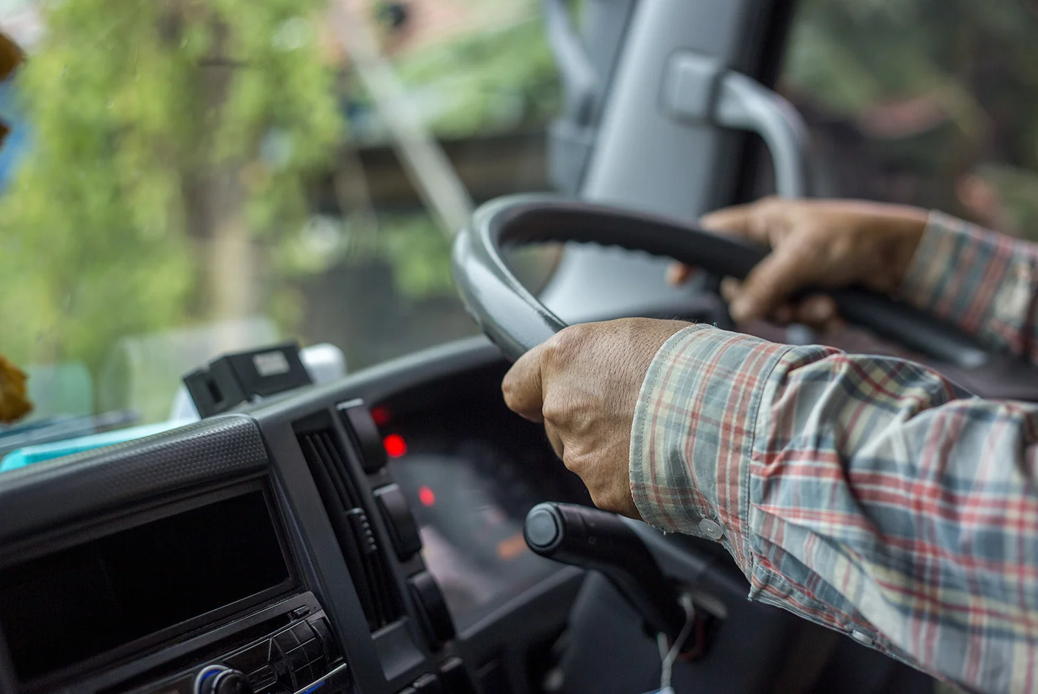 Close-up of a person’s hands gripping a steering wheel inside a truck cab, with dashboard controls and blurred greenery visible through the windshield.