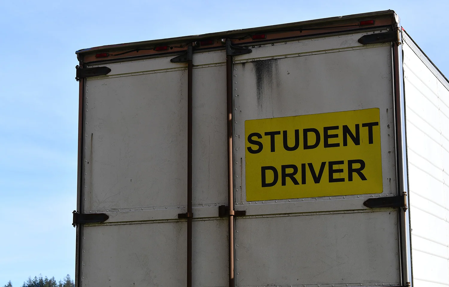 Rear view of a large white truck with a bright yellow sign that reads “STUDENT DRIVER” in bold black letters.