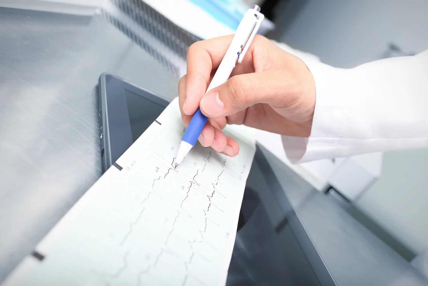 A clinician reviewing and marking an EKG printout with a pen on a stainless‑steel surface.