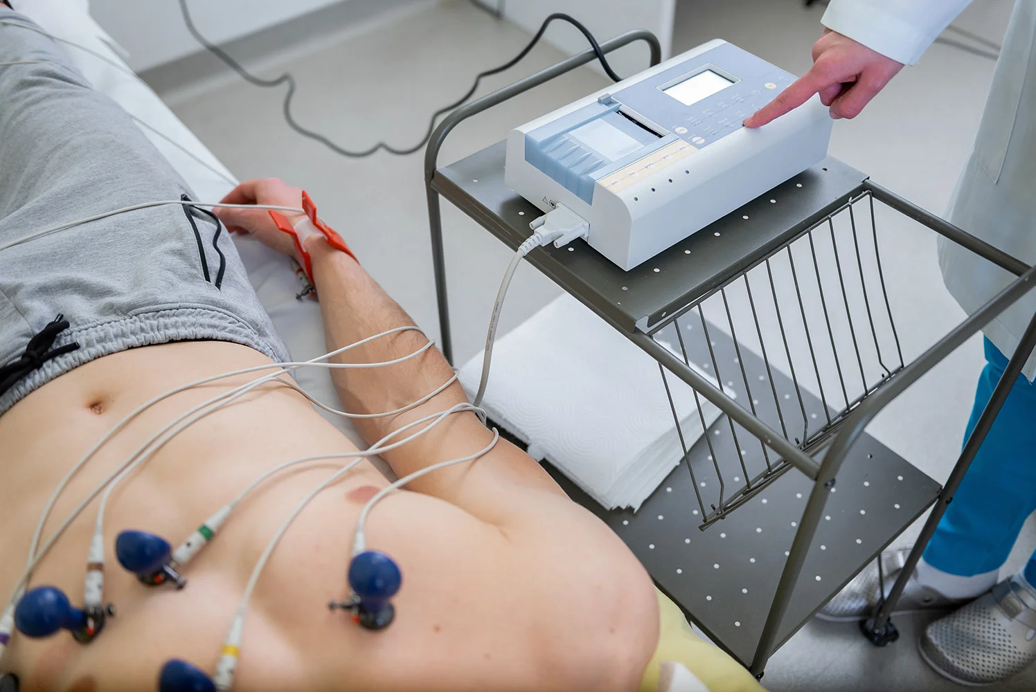 A patient lying on an exam table with EKG electrodes attached to their chest while a healthcare provider selects settings on the EKG machine.