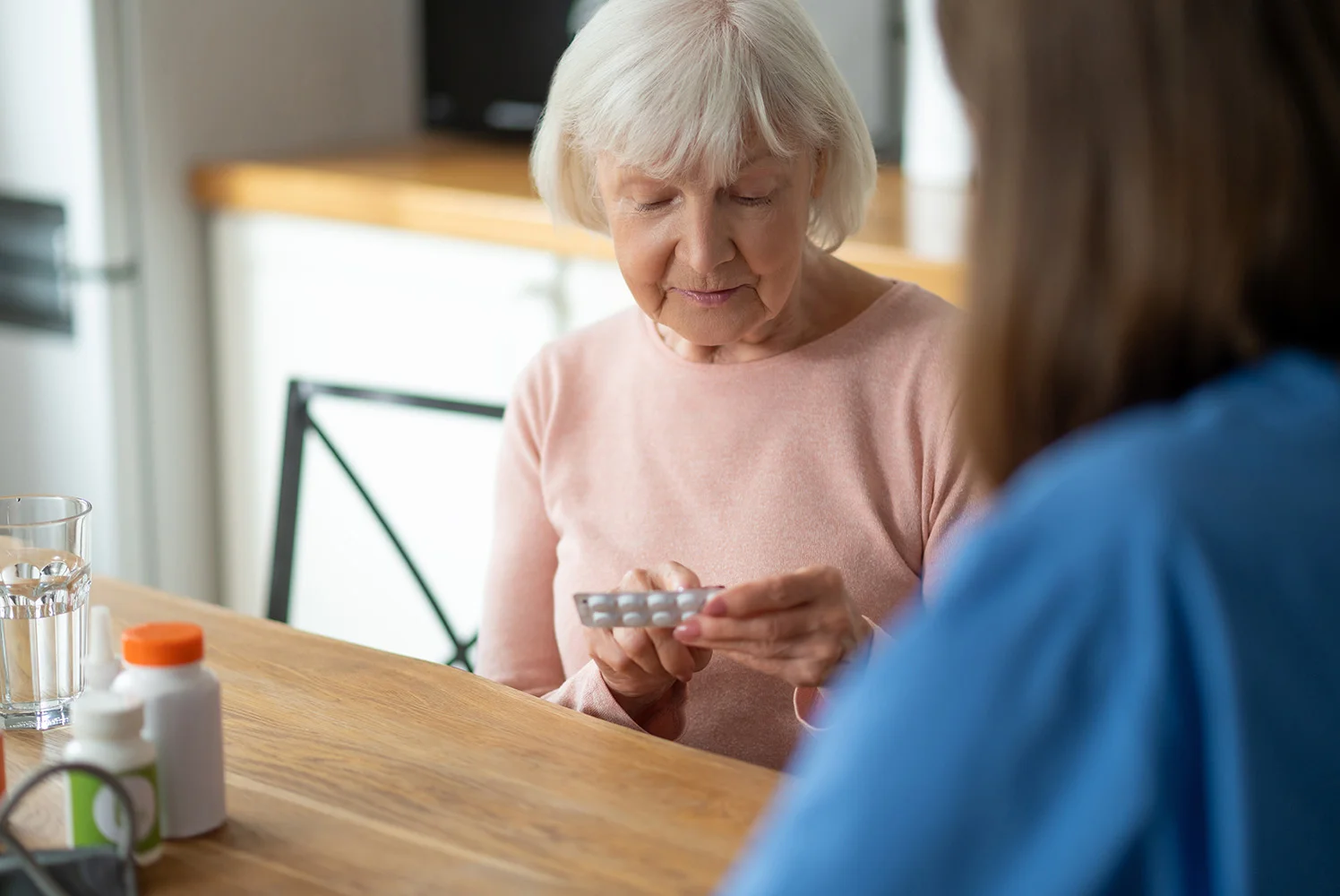 An older adult sitting at a table holding a blister pack of medication, with various pill bottles and a glass of water nearby.