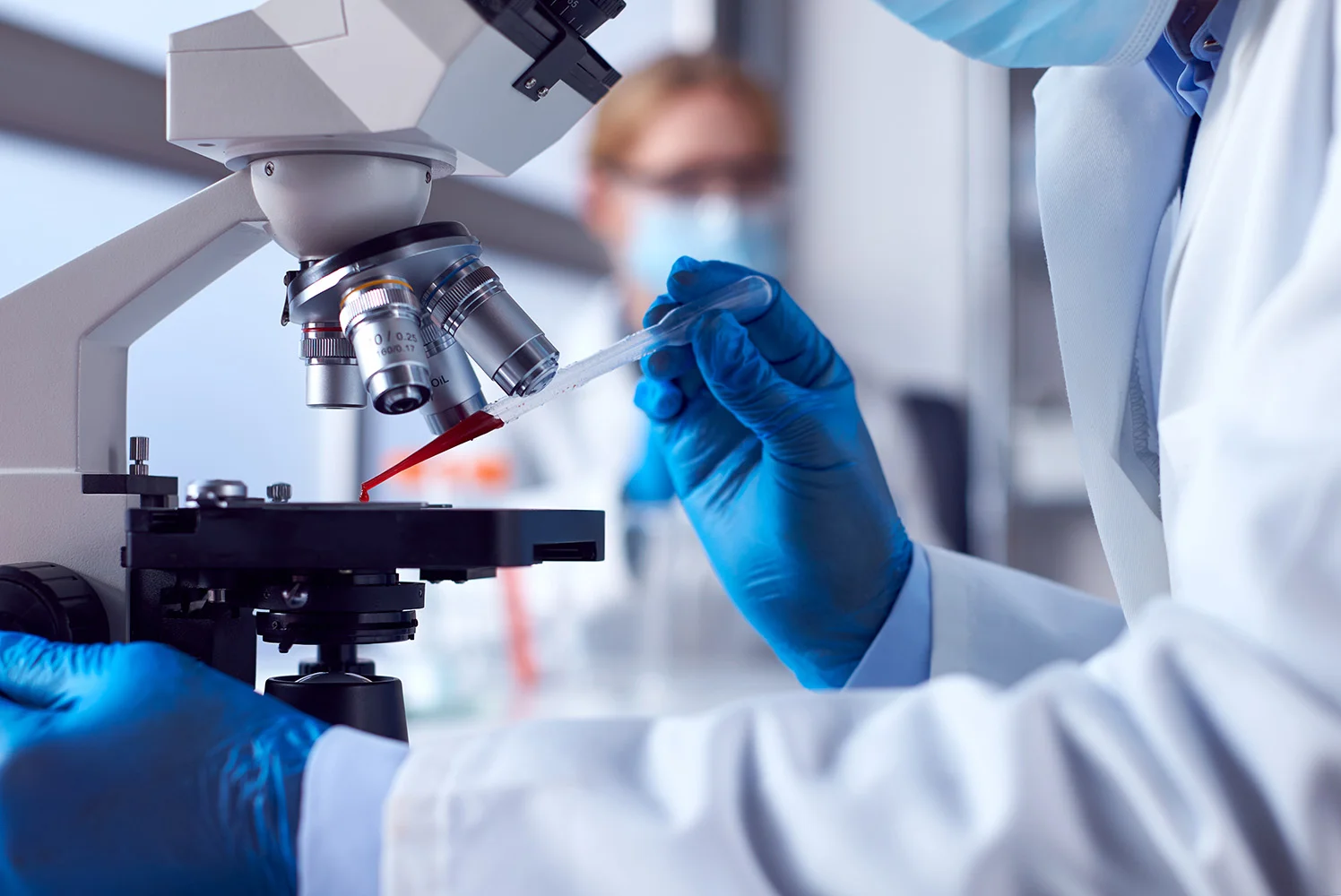 Laboratory technician using a pipette to place a sample under a microscope, wearing protective gloves and a lab coat in a clinical setting.