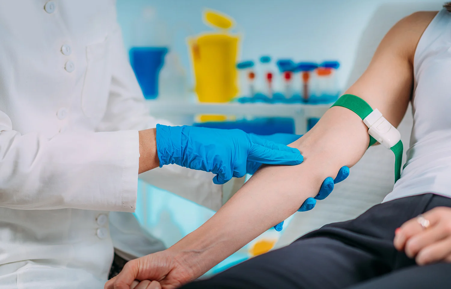 Healthcare professional wearing blue gloves preparing a patient’s arm for a blood draw, with a green tourniquet applied and medical supplies visible in the background.