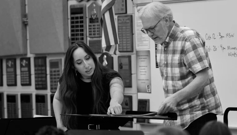 Clare Howard stands beside another person at a music stand, reviewing sheet music in a band rehearsal room filled with musicians, plaques, and a whiteboard.