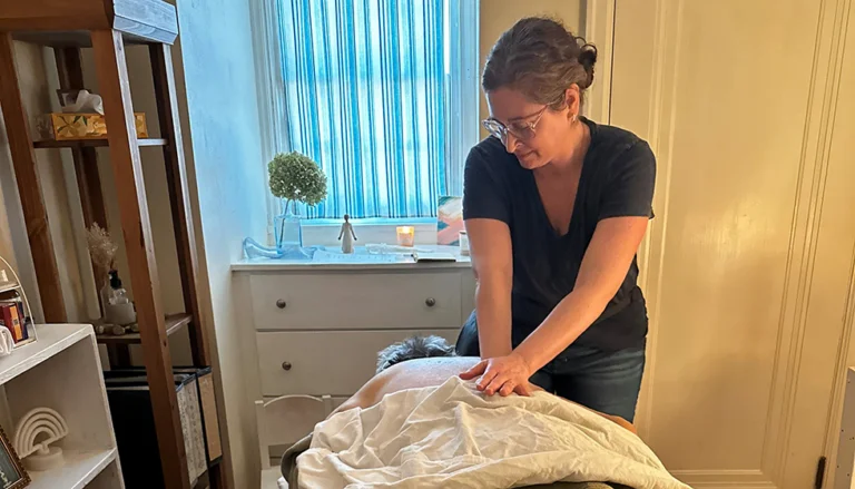 Lindsay Bejblik performs a massage in a softly lit room with a massage table, shelving, and a small dresser topped with a candle and decorative plant near a window.