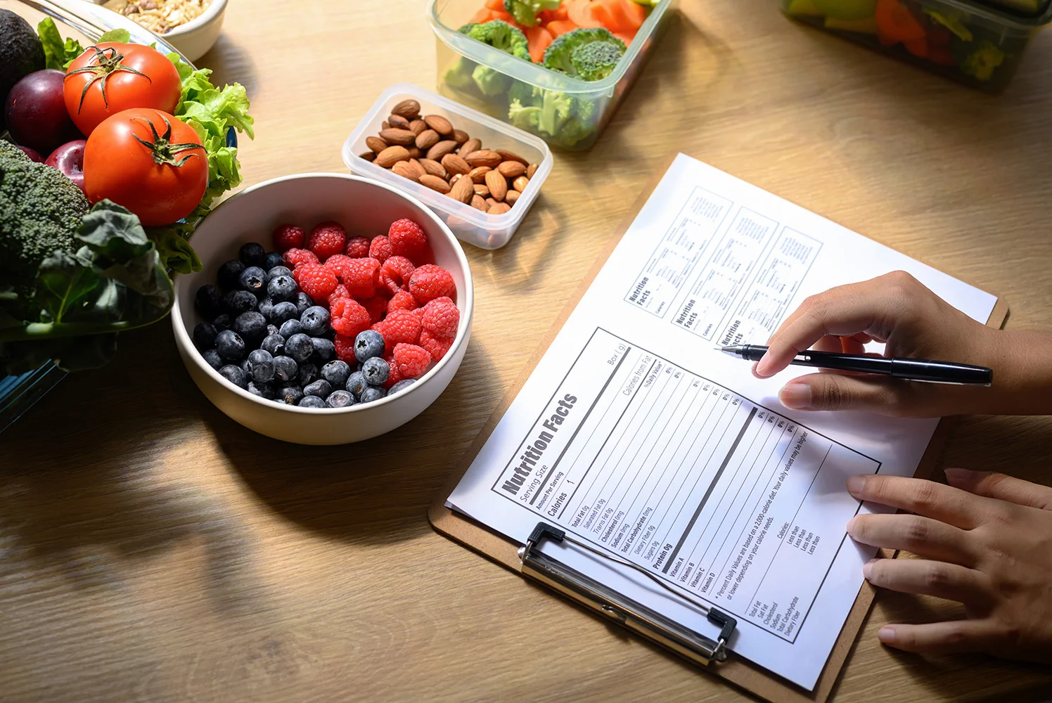 Fresh fruits and vegetables, nuts, and meal prep containers on a table beside a clipboard with a nutrition plan being filled out by hand