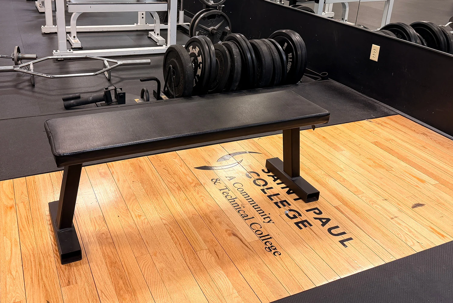 Weight bench in a gym with barbells and plates behind it on a floor labeled with Saint Paul College branding