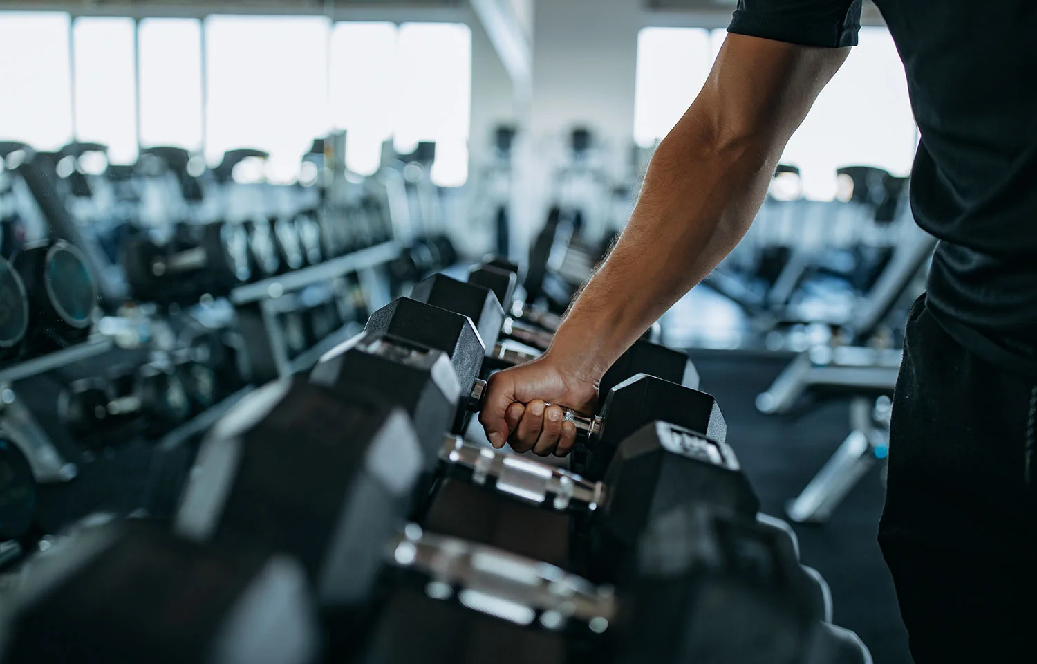 Person reaching for a dumbbell from a rack in a well-lit gym filled with weight equipment.