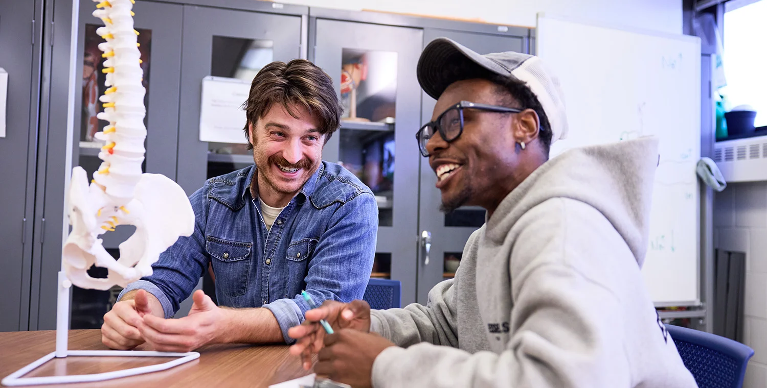 A person in a denim shirt and another person in a hoodie sit at a table in a classroom or lab, discussing a clipboard while a model of a human spine is displayed beside them.