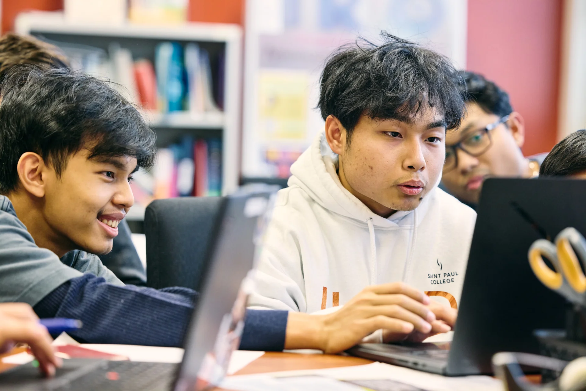 Students gathered around laptops in a classroom, collaborating and smiling while working on a project.