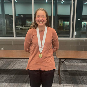A SkillsUSA gold medalist smiles while standing indoors, wearing a medal around their neck.