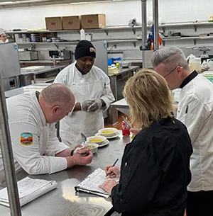 Culinary Arts competitors work at stainless steel prep tables while judges observe and take notes during a SkillsUSA competition.